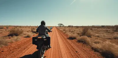 Motorcyclist riding along a red dirt road through the Northern Territory outback under a clear blue sky.