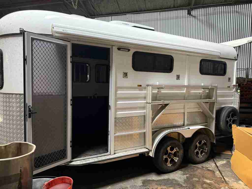 White tandem-axle horse float with an open side door and awning parked inside a shed.